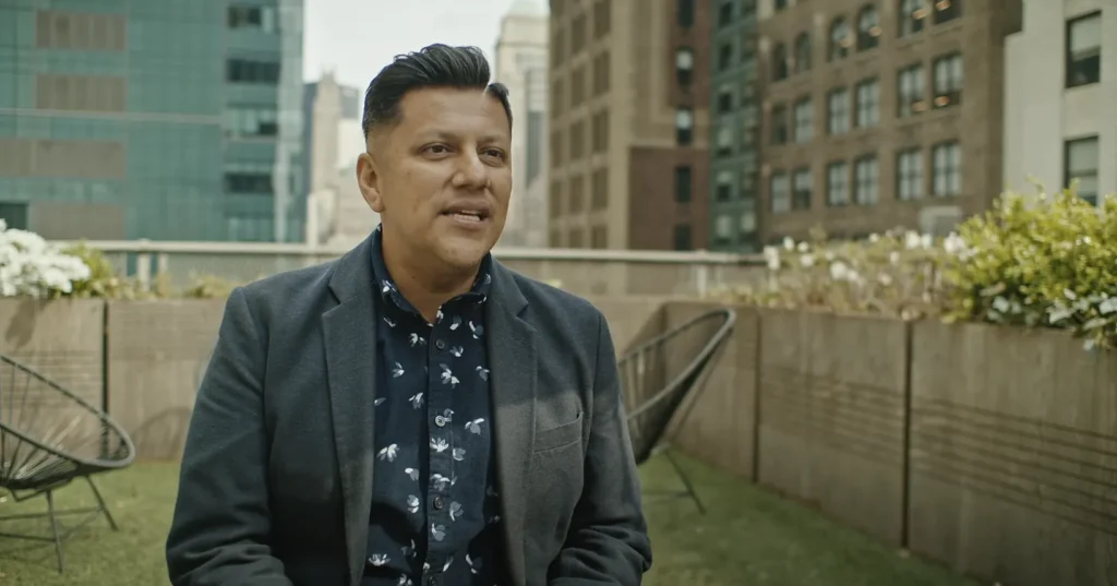 A man is being interviewed on a rooftop with a city skyline in the background.